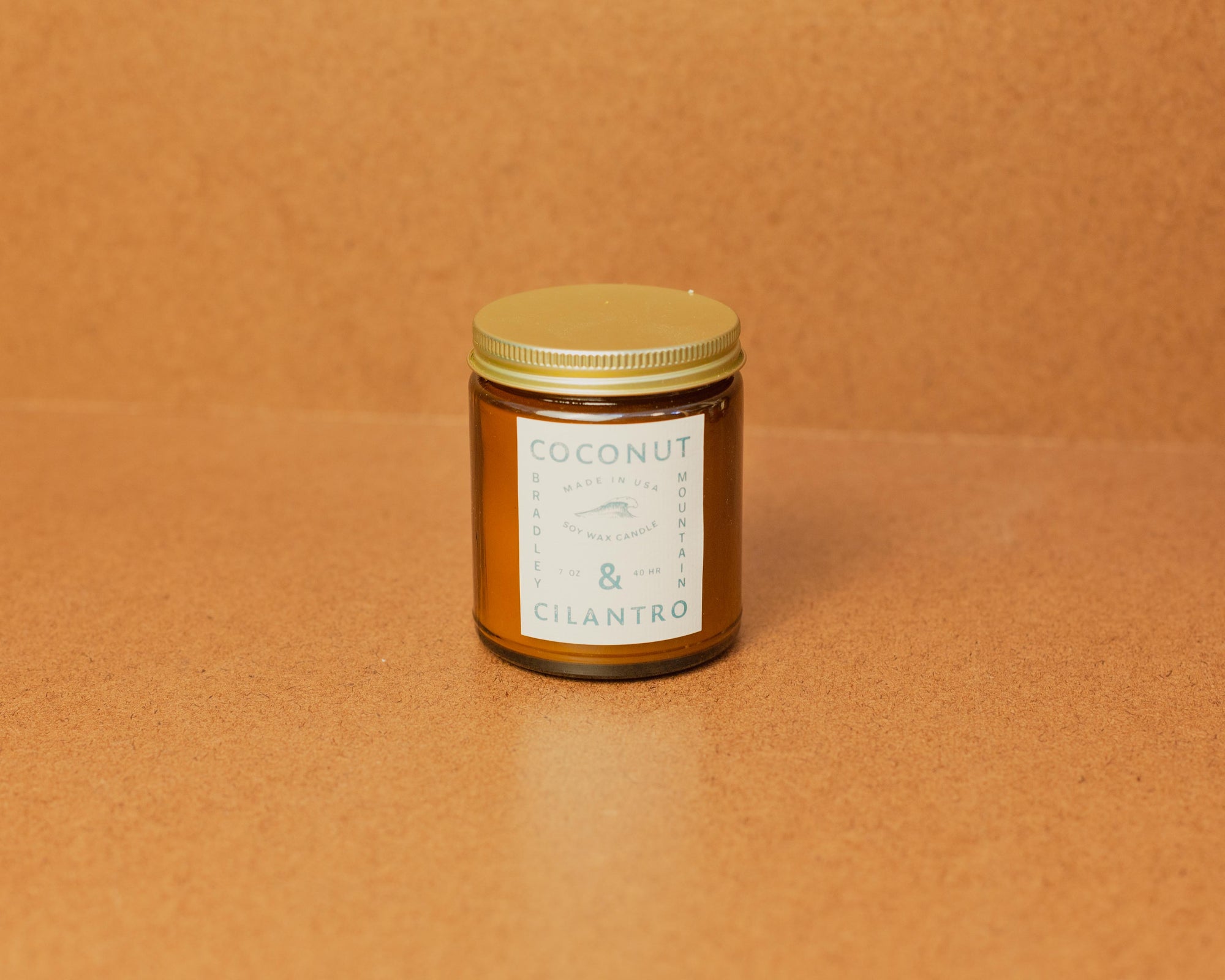 A small amber glass jar with a lid, labeled 'Coconut' and 'Cilantro', containing a candle. The jar is placed on a brown surface.