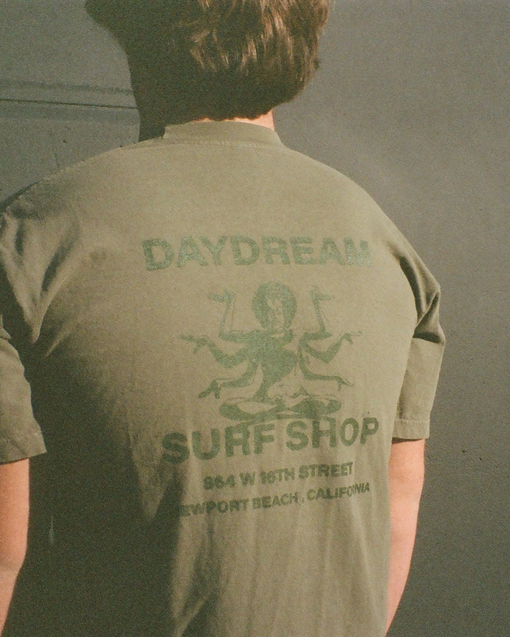 Person wearing a green t-shirt with 'Daydream Surf Shop' printed on the back, standing against a dark background.