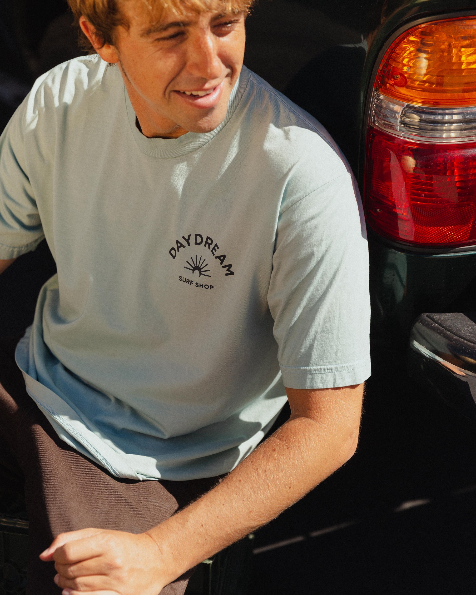 Person wearing a light blue t-shirt with 'Daydream Surf Shop' logo sitting next to a car.