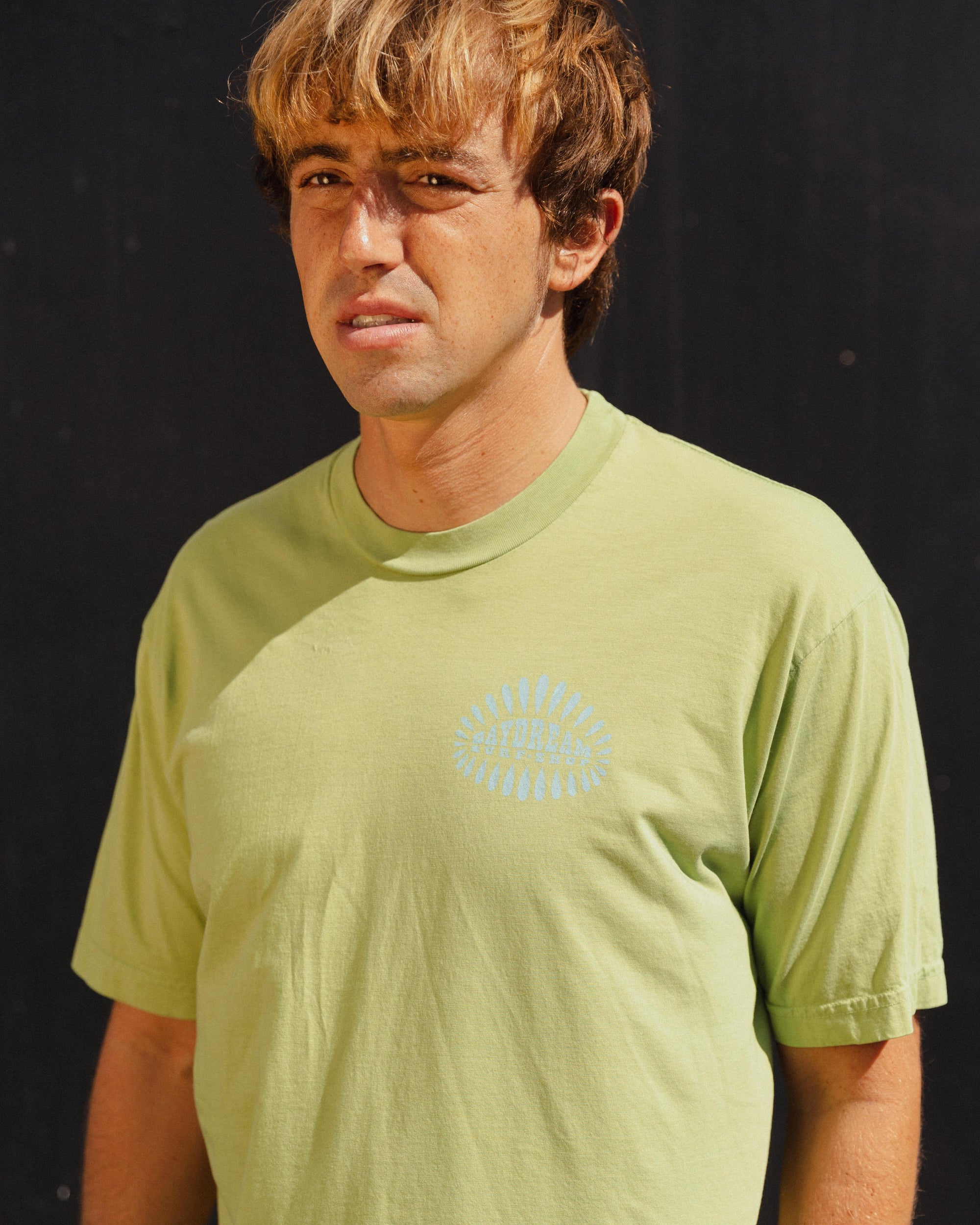 Person wearing a light green t-shirt with a blue 'Daydream Surf Shop' logo against a black background